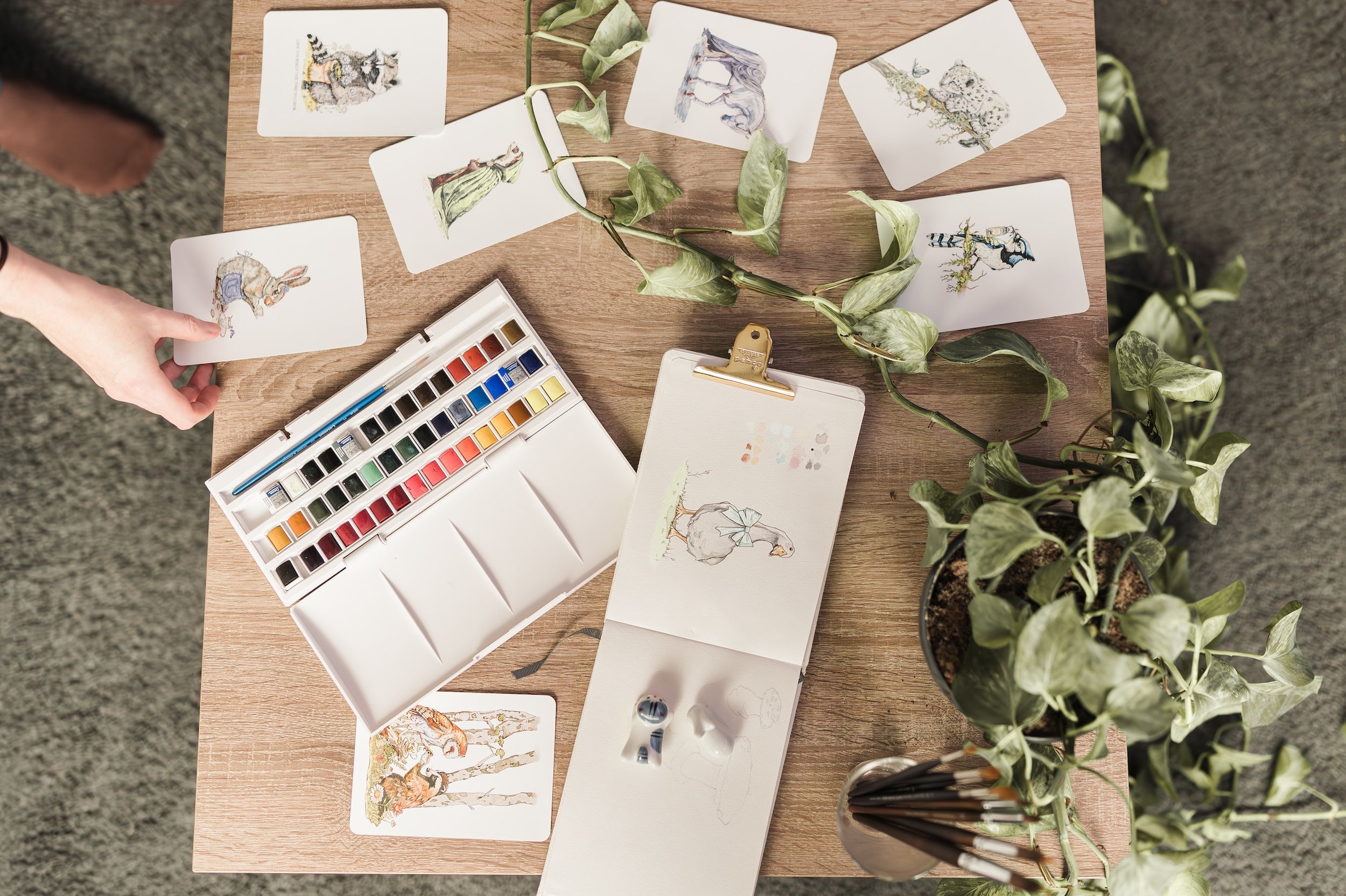 Julia Hope’s table displaying her hand-painted watercolor art cards, whimsical animal illustrations, and nature-inspired artwork, with her hand arranging the pieces.